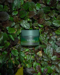 A frosted green cosmetic jar with the brand name 'VATYAI' printed in white, nestled among dense green and burgundy foliage.